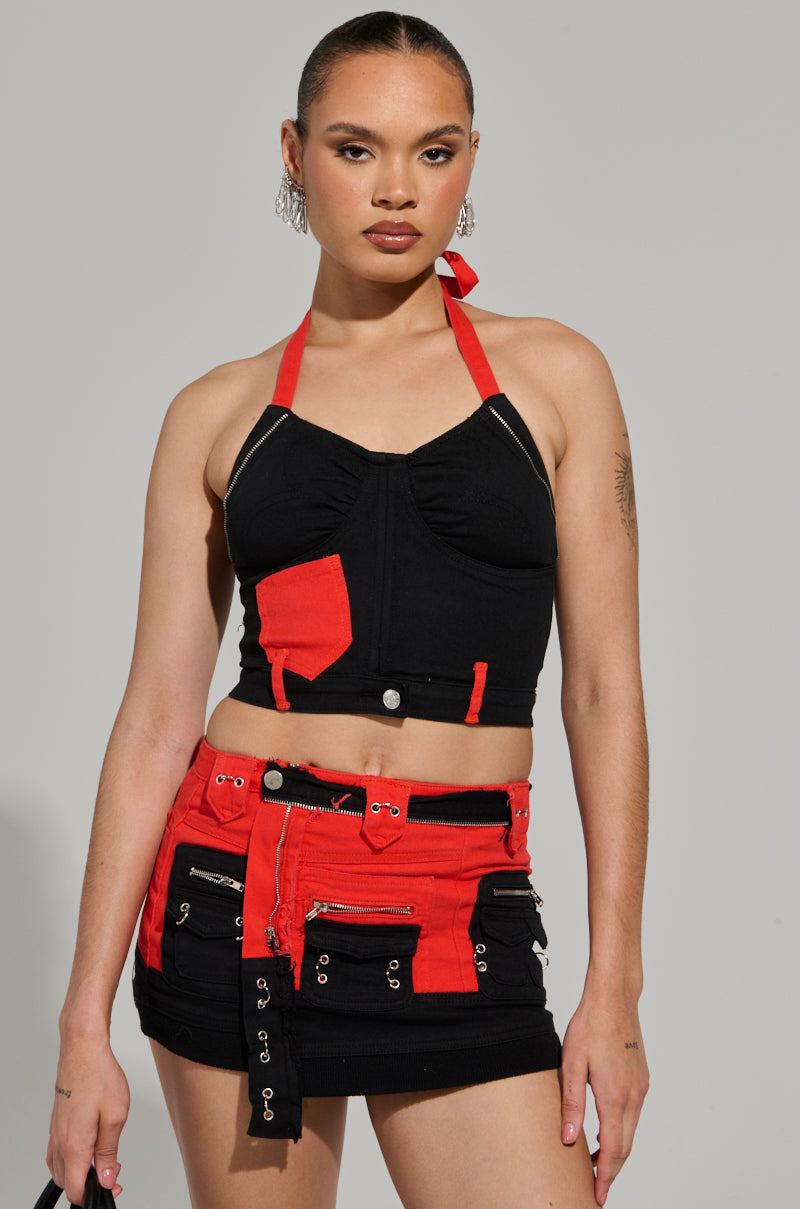 A person is posing in a MOTOR SPORT CORSET TOP IN BLACK & RED with a mini skirt that has multiple pockets and decorative zippers. Their hair is pulled back, highlighting statement earrings against a neutral background.