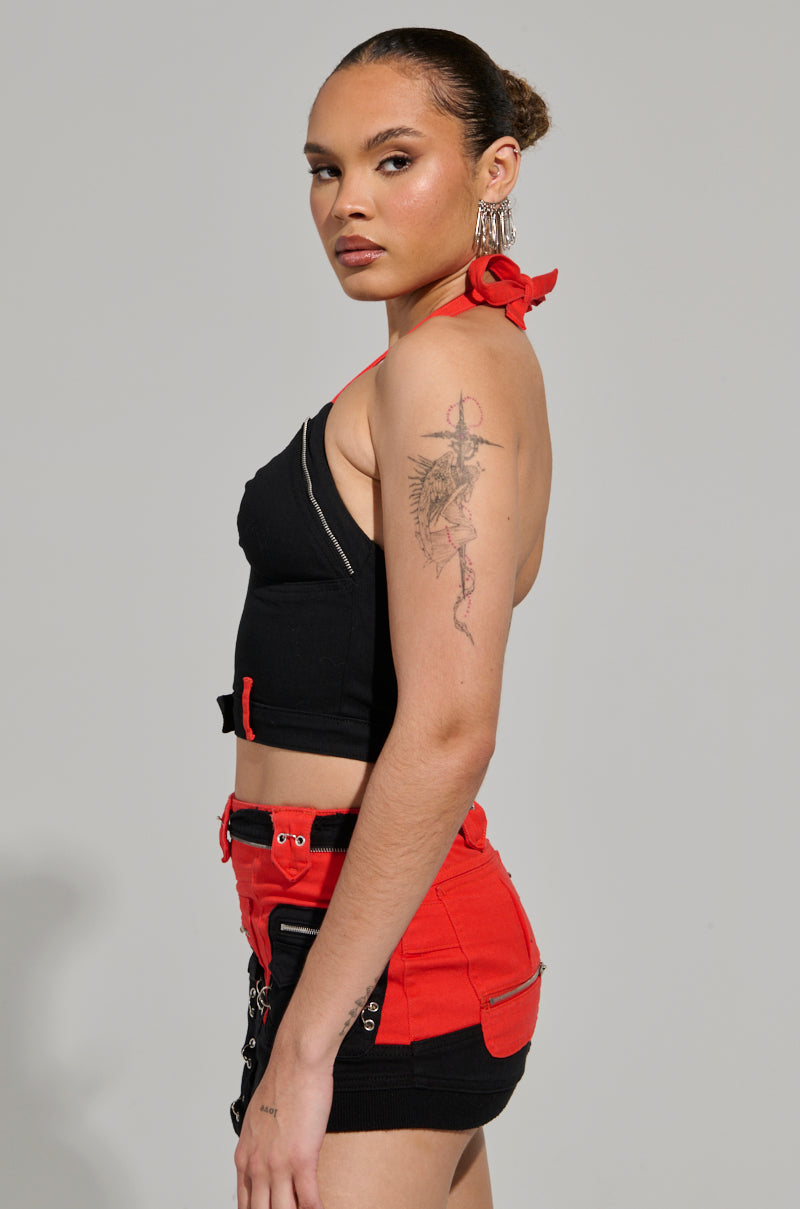 A person wearing the Motor Sport Corset Top in Black & Red and red belted shorts with a color-block design stands in profile. They have their hair tied up, sport large hoop earrings, and showcase a tattoo on their left arm.