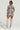 A person stands confidently in an AKIRA Label Flower Child floral camo jacket paired with the FLOWER CHILD PRINTED MINI SKIRT. They accessorize with oversized sunglasses, large earrings, and knee-high beige boots. The plain background highlights the fashionable ensemble.