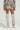 A person dressed in the FLOWER CHILD PRINTED MINI SKIRT from the AKIRA Label, along with a matching floral camo jacket ensemble and gray over-the-knee boots, posed against a simple, light-colored background.