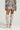 A person wearing an AKIRA Label Flower Child floral camo jacket with matching shorts and a light beige over-the-knee boots stands against a plain background, complemented by the FLOWER CHILD PRINTED MINI SKIRT.