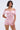 A woman with a bob haircut models the LA LA LOVE ME SATIN ROMPER, an off-the-shoulder, short-sleeve pink satin romper, standing with one arm behind her back against a light backdrop.