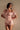 Wearing the LA LA LOVE ME SATIN ROMPER, a light pink off-the-shoulder satin romper with short puff sleeves and a matching neck ribbon, a woman stands against a neutral background, gazing slightly to the side.