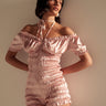 Wearing the LA LA LOVE ME SATIN ROMPER, a light pink off-the-shoulder satin romper with short puff sleeves and a matching neck ribbon, a woman stands against a neutral background, gazing slightly to the side.