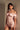 A woman with short dark hair and statement earrings stands against a light wall, wearing the LA LA LOVE ME SATIN ROMPER—a shiny, pale pink romper with puff sleeves—while looking at the camera.