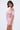 A woman wearing a shiny, light pink, off-the-shoulder mini dress similar to the LA LA LOVE ME SATIN ROMPER and large pearl earrings stands against a plain background, gazing at the camera with her head softly tilted.