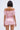 A woman with short dark hair is seen from behind, wearing the LA LA LOVE ME SATIN ROMPER in shiny light pink with ruffled details and subtle puff sleeves, standing against a plain light background.