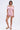 A woman with short hair and pearl earrings poses against a plain background in the LA LA LOVE ME SATIN ROMPER, a shiny light pink satin one-piece with off-the-shoulder puff sleeves. She wears beige slide sandals, one hand by her side and the other on her thigh.