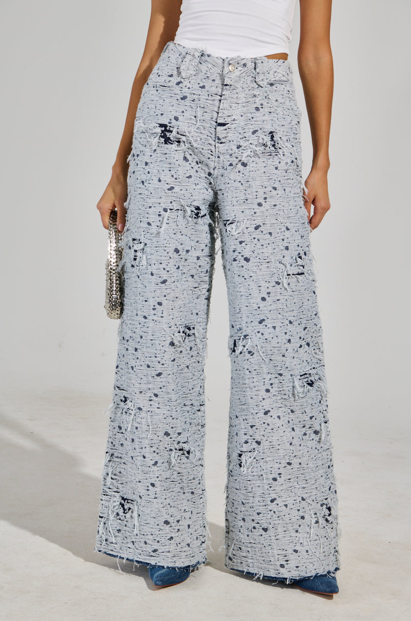 Wearing the NEVER LOSE ME RELAXED DISTRESSED DENIM, featuring a high-rise waist and wide-leg silhouette, a person pairs their outfit with a white top and holds a small silver handbag while standing gracefully against a plain background.