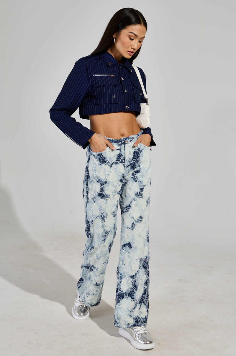 A person with long dark hair wears a navy pinstripe cropped jacket and light, patterned "Too Cool For You Denim Pant" in a wide-leg silhouette, paired with silver sneakers. They hold a white bag and walk on a plain background, exuding an effortlessly cool vibe.