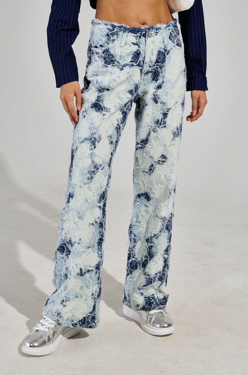 A person is wearing the TOO COOL FOR YOU DENIM PANT, featuring high-waisted, light blue jeans adorned with a floral pattern. They are also sporting a navy blue cropped sweater and white sneakers with metallic silver toes. The plain light gray backdrop accentuates the outfit effortlessly.