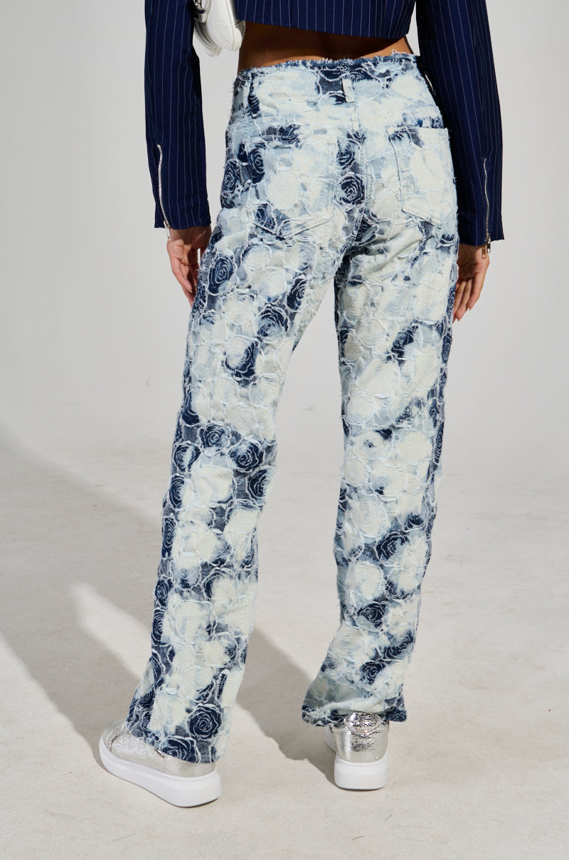 A rear view of a person sporting the "TOO COOL FOR YOU DENIM PANT," featuring blue and white floral patterns, paired with white sneakers adorned with silver glitter heels. Their ensemble is completed by a dark pinstripe jacket against a plain, light-colored background.