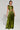 A person wearing a shiny green sleeveless top and the METALLIC DREAM PANT with a wide-leg silhouette stands against a plain background. They hold a phone, have short hair, and are accessorized with bracelets and visible tattoos on their stomach and arms.