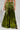 A person wearing the METALLIC DREAM PANT in a shimmering metallic green color, featuring a wide-leg silhouette and high-rise waist, holds a small white clutch. The outfit is complemented by black shoes and silver bracelets, with a tattoo visible on their waist against a plain light gray background.