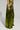 A person wearing the METALLIC DREAM PANT in a shiny, metallic green with wide legs and a high-rise waist stands against a plain background. The pants reflect light, creating a glamorous appearance. The individual's hand, adorned with bracelets, rests casually by their side.