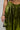 A person dressed in METALLIC DREAM PANT, featuring a shiny green metallic finish with a wide leg silhouette, is holding a small silver purse. A tattoo and navel piercing are visible on their midriff against a plain background.