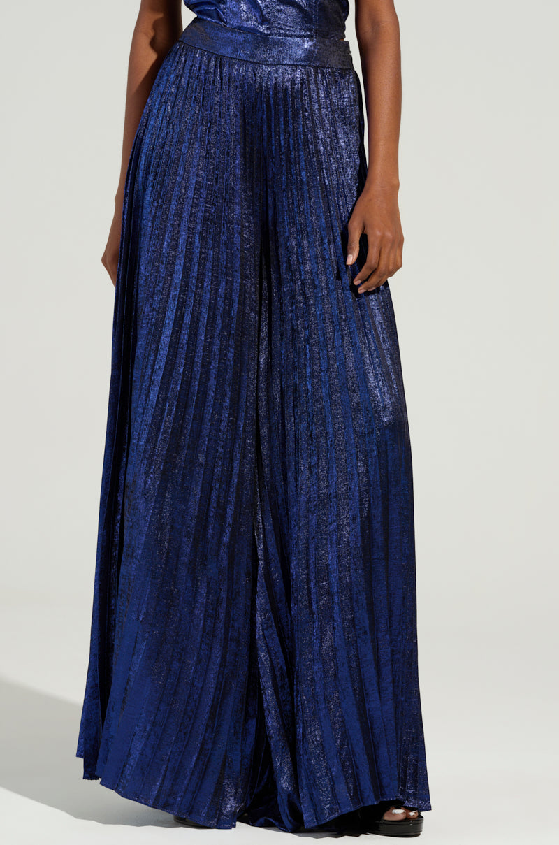 A person wearing the TAKE MY HAND PANT in shiny blue, featuring a high-rise, pleated wide-leg design, with their hand resting on the side against a light gray background.