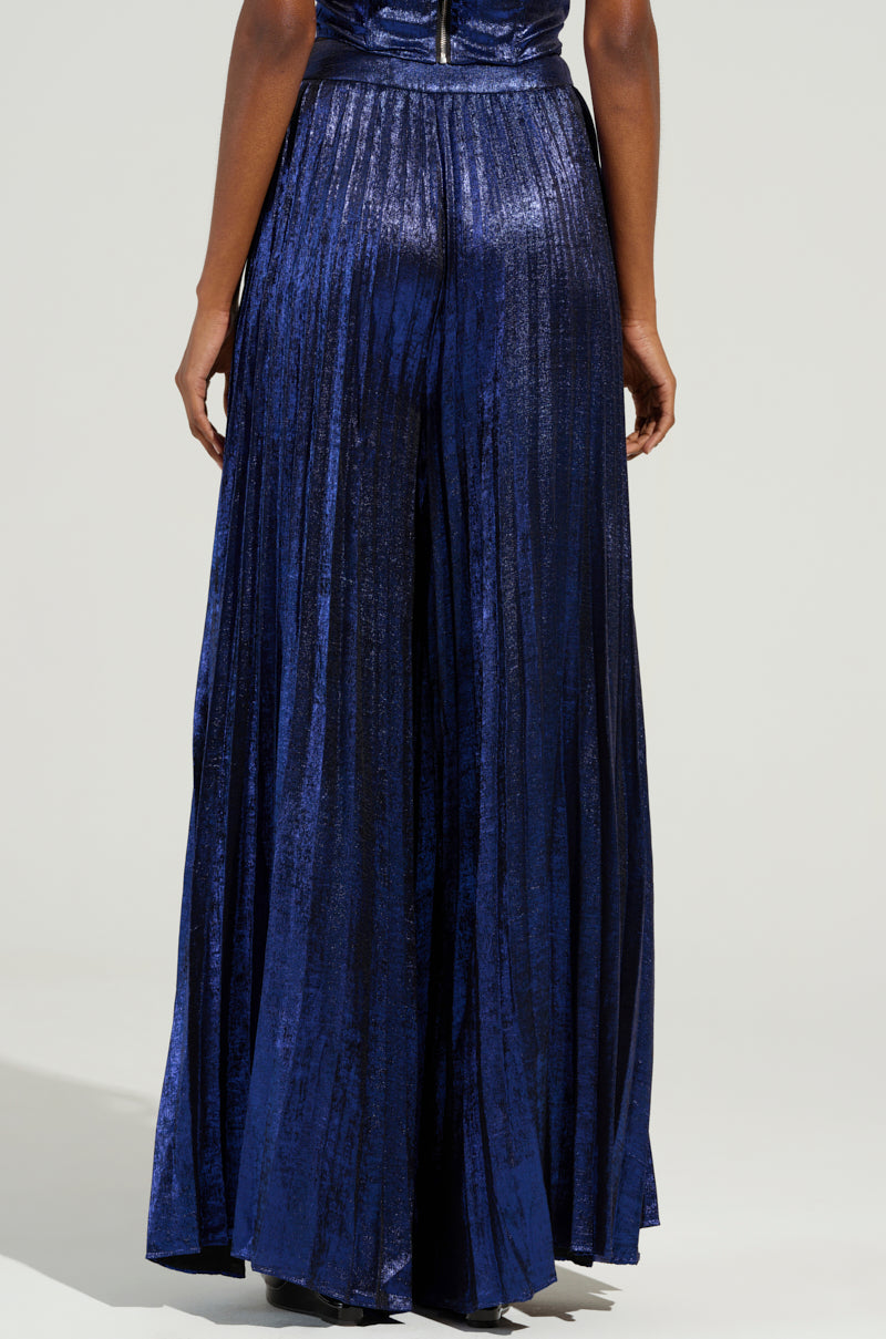 Rear view of a person wearing the shimmering TAKE MY HAND PANT in deep blue, standing against a neutral background. The fabric reflects light, highlighting its texture and elegant flow reminiscent of a pleated gown silhouette.