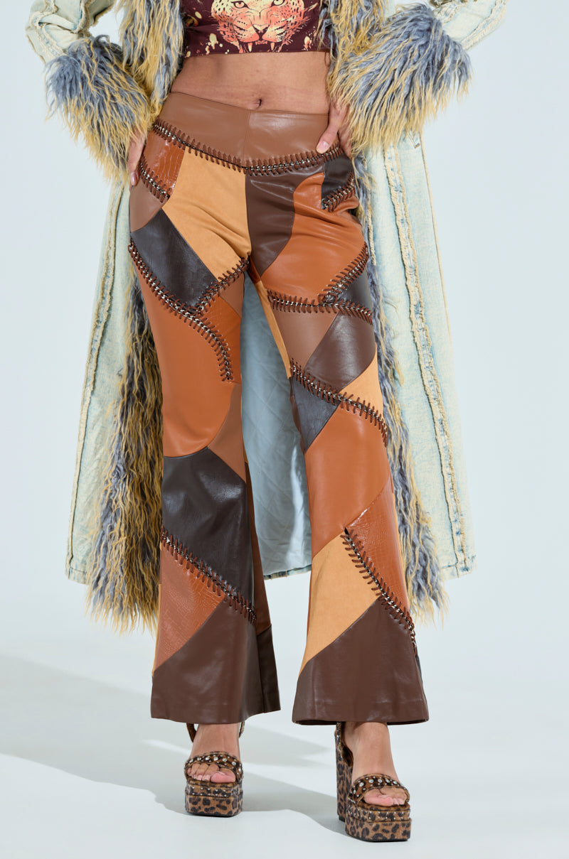 A person wearing TALK LATER PATCH FAUX LEATHER FLARE PANT in brown with zigzag stitches has their hands in the pockets. Completing the look is a long, fur-trimmed coat and animal print platform sandals. 2