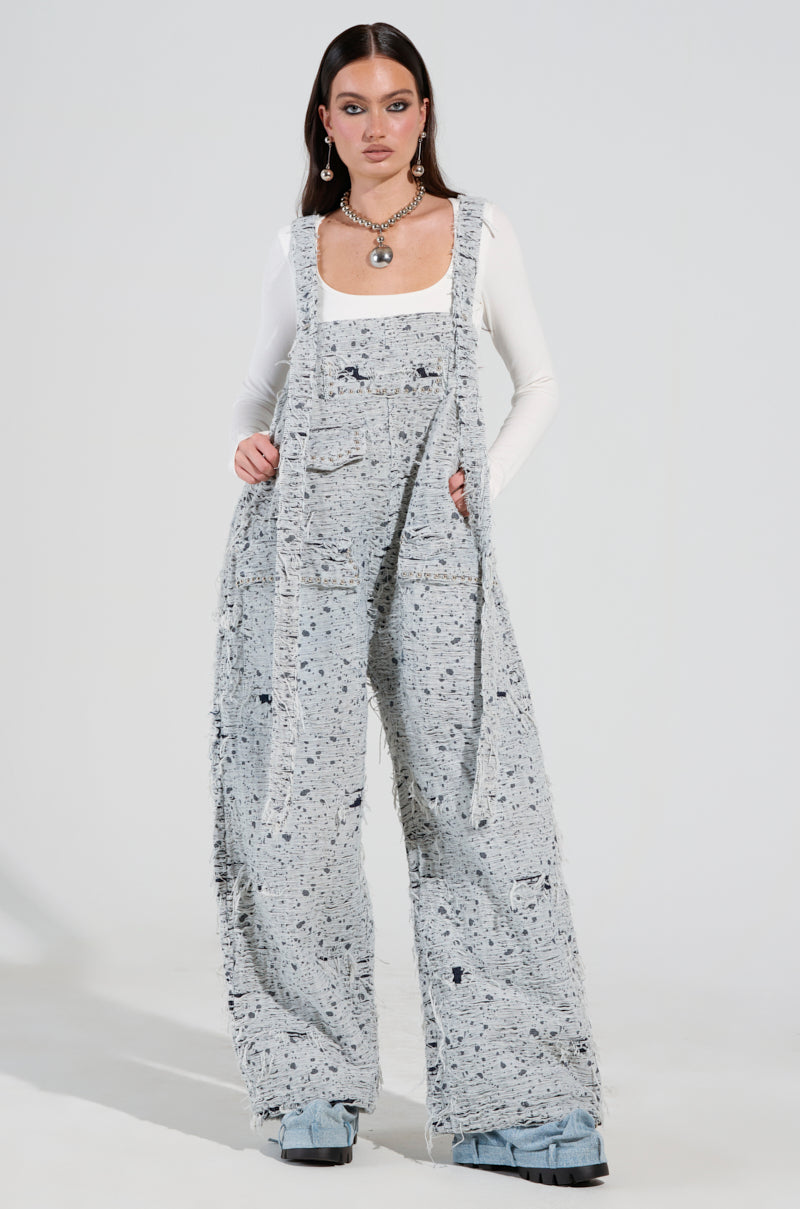 A person confidently stands wearing the HERE TO SHRED DISTRESSED DENIM OVERALLS, which feature a textured light blue fabric with wide-leg pants. They pair it with a white long-sleeve top and statement jewelry, while their long, dark hair complements the chic ensemble against a plain, well-lit backdrop.
