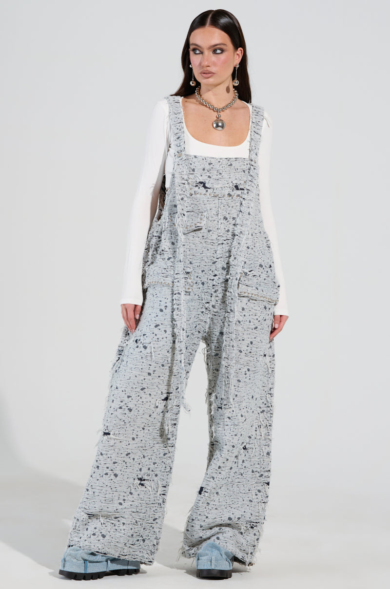 A person confidently stands in HERE TO SHRED DISTRESSED DENIM OVERALLS in a gray speckled style, paired with light blue clogs. The outfit is layered over a white long-sleeve top, and accessorized with a chunky necklace and long earrings, all set against a plain white background.