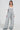 A person confidently stands in HERE TO SHRED DISTRESSED DENIM OVERALLS in a gray speckled style, paired with light blue clogs. The outfit is layered over a white long-sleeve top, and accessorized with a chunky necklace and long earrings, all set against a plain white background.