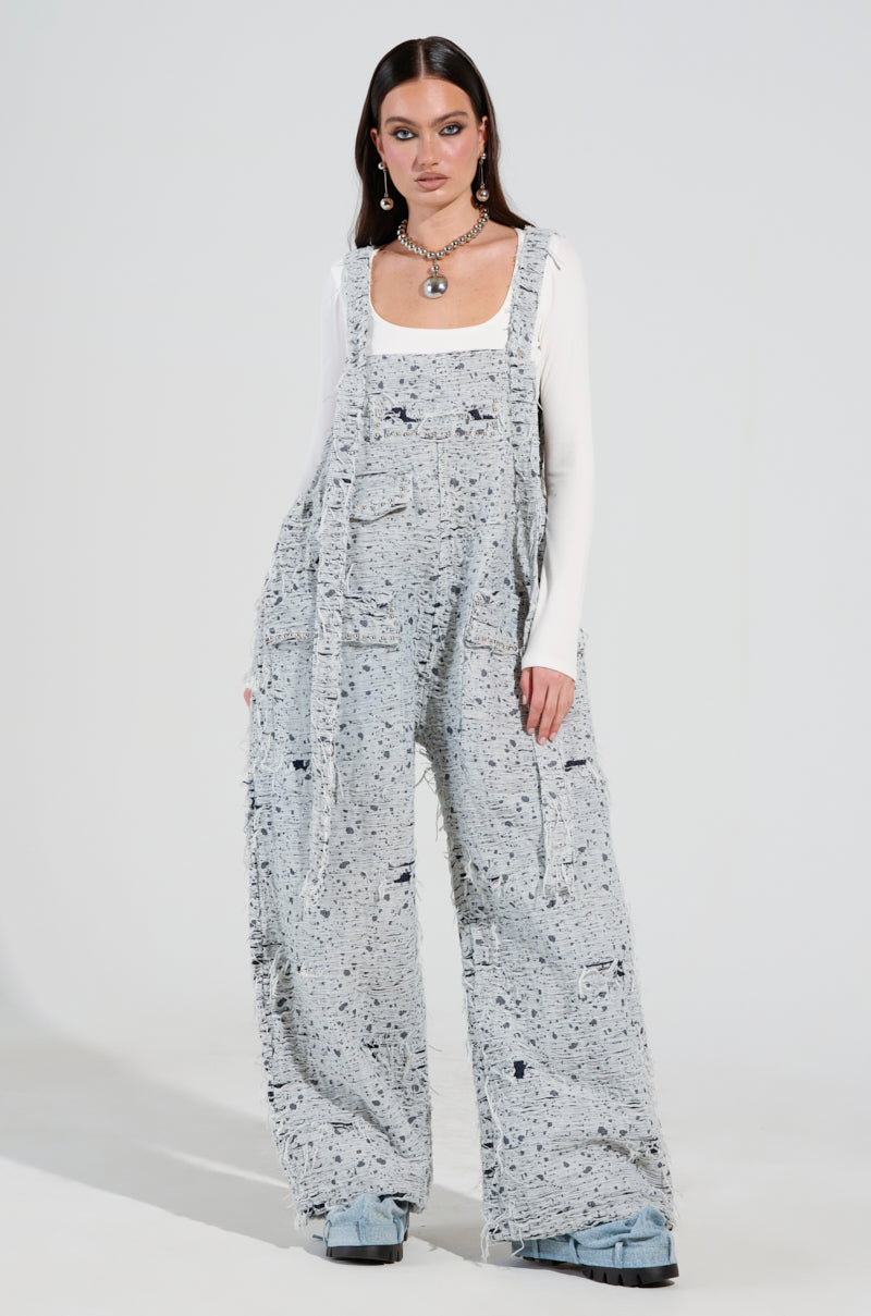 A person wearing the HERE TO SHRED DISTRESSED DENIM OVERALLS paired with a white long-sleeve top and chunky shoes stands against a plain background. They accessorize with a layered necklace and hoop earrings, adding a touch of flair to the wide-legged ensemble.
