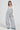 A person wearing the HERE TO SHRED DISTRESSED DENIM OVERALLS paired with a white long-sleeve top and chunky shoes stands against a plain background. They accessorize with a layered necklace and hoop earrings, adding a touch of flair to the wide-legged ensemble.