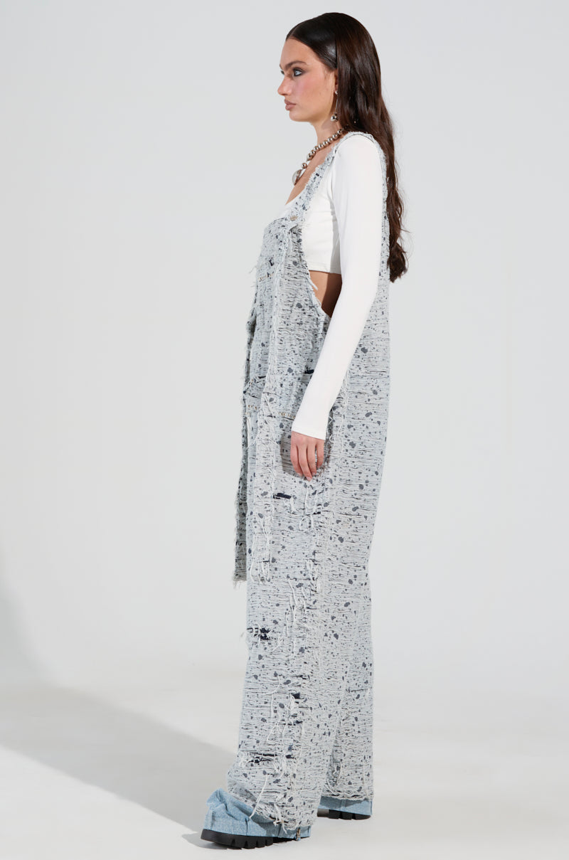 A woman modeling the "Here to Shred Distressed Denim Overalls," featuring stud-embellished pockets, stands in profile. She has long, dark hair and pairs her outfit with a white long-sleeve top and light blue shoes, set against a plain backdrop.