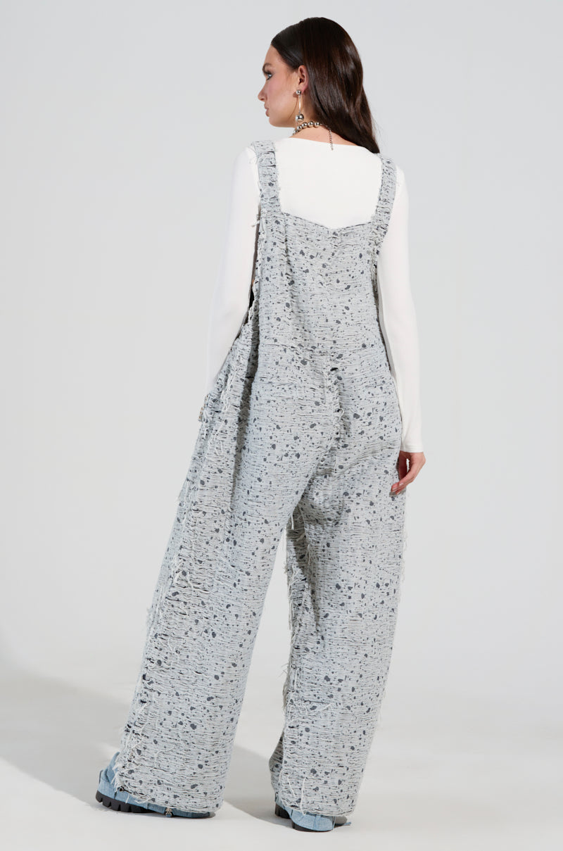 A person with long brown hair is seen from behind, wearing the HERE TO SHRED DISTRESSED DENIM OVERALLS with stud-embellished pockets over a white long-sleeve top. They complement their outfit with statement earrings and blue shoes, all set against a plain background.