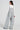 A person with long brown hair is seen from behind, wearing the HERE TO SHRED DISTRESSED DENIM OVERALLS with stud-embellished pockets over a white long-sleeve top. They complement their outfit with statement earrings and blue shoes, all set against a plain background.