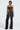 A person confidently stands against a light background dressed in the DANCING THE NIGHT AWAY WOVEN RHINESTONE JUMPSUIT, which boasts a stylish strapless design with long silver fringe detailing and a wide-leg silhouette, paired elegantly with silver heels.
