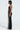 A person stands in profile, wearing the "Dancing the Night Away" woven rhinestone jumpsuit, featuring a strapless design with long silver fringe embellishments cascading down the front. Their shoulder-length dark hair is accentuated against a plain light background.