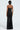 A person stands with their back to the camera, highlighting the strapless style of the DANCING THE NIGHT AWAY WOVEN RHINESTONE JUMPSUIT in black. This jumpsuit is embellished with rhinestones, featuring wide-leg pants and a fitted top, finished with a zipper down the back against a simple, light backdrop.