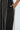 A close-up image captures a person in the Dancing The Night Away Woven Rhinestone Jumpsuit, showcasing its black wide-leg silhouette adorned with vertical rows of sparkling rhinestones. Their hand rests casually in the pocket, while the neutral background accentuates the shimmering details.