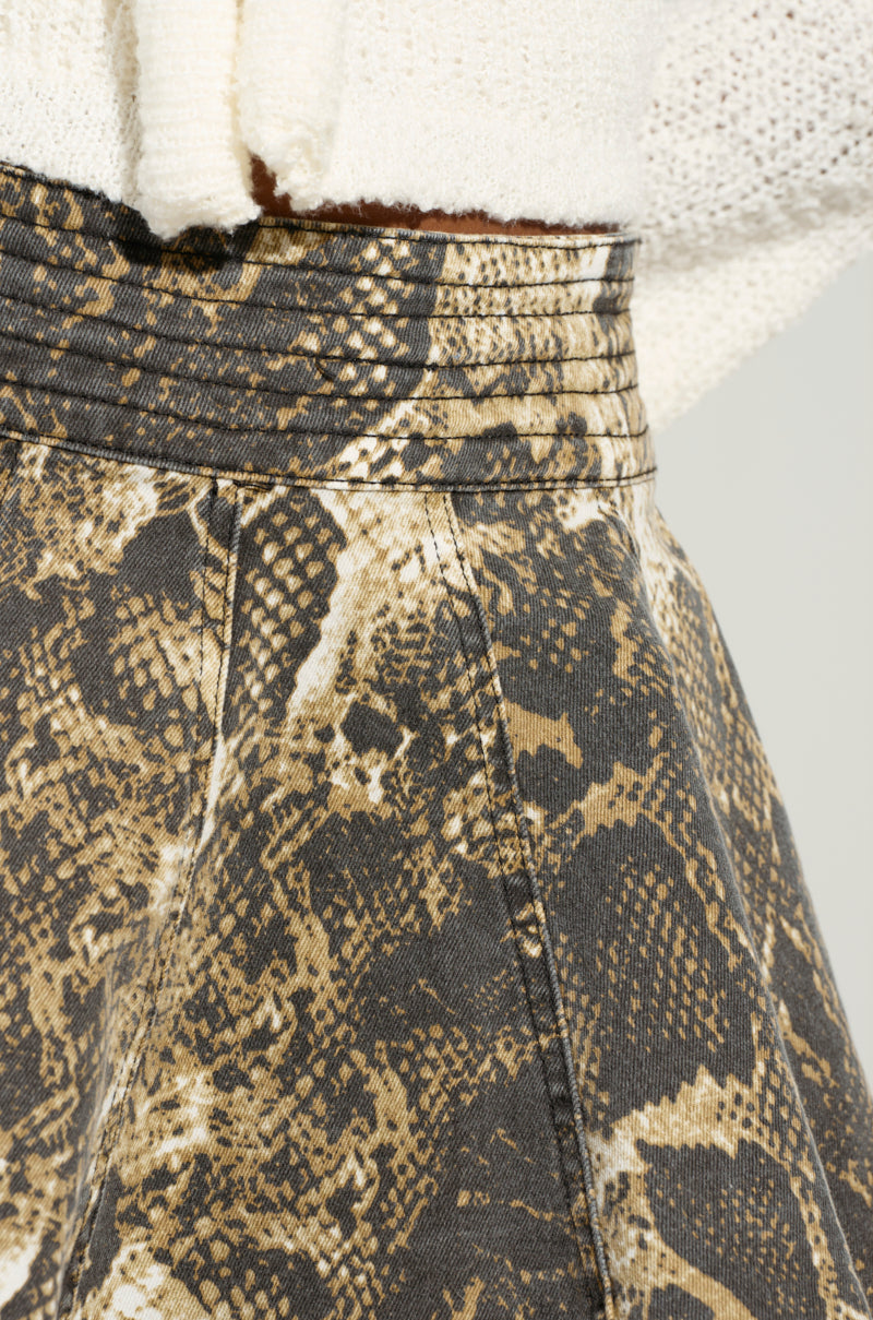 Close-up of a person's waist adorned in the WORST INTENTIONS MINI SKIRT with a brown and beige snakeskin pattern, perfectly complemented by a tucked-in cream sweater. The skirt features a high-rise fit with its intricate design, while the textured sweater adds an extra layer of style.