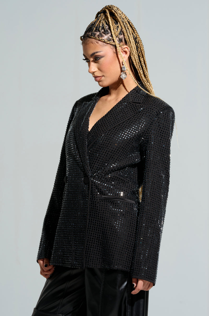 A person with long braided hair wearing the BOTTOMS UP EMBELLISHED BLAZER, adorned with rhinestones, and black pants stands against a plain background, looking to the side.