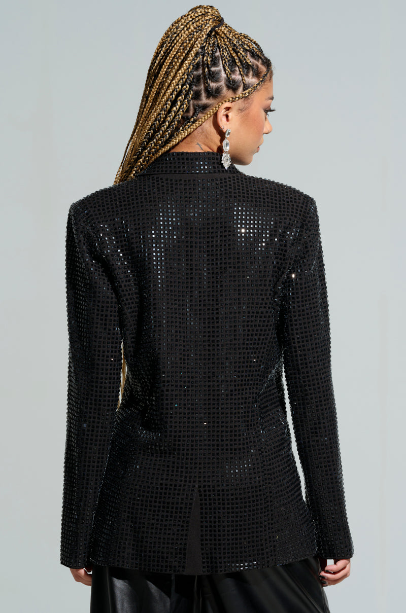 A person with long braided hair is wearing the BOTTOMS UP EMBELLISHED BLAZER, which sparkles in black and features square embellishments and rhinestones. They are facing away from the camera, highlighting the intricate details of this embellished garment against a neutral light gray background.