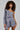 A person with curly hair is wearing the stylish OUT OF OFFICE BLAZER ROMPER, featuring an asymmetrical design and a gray one-sleeve silhouette, accessorized with layered necklaces. They confidently pose against a plain background, exuding a business-chic flair.