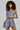 A woman with curly hair is donning the stylish OUT OF OFFICE BLAZER ROMPER, featuring an asymmetrical design with one shoulder covered, projecting a business-chic vibe. Her confident expression is complemented by a layered necklace that seamlessly blends elegance and modernity.