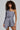 A person with curly hair wearing the distinctive OUT OF OFFICE BLAZER ROMPER in a grey asymmetrical one-sleeve design stands confidently against a plain background, accessorized with a layered necklace, epitomizing business-chic style.