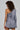 A person with curly hair is wearing the business-chic "OUT OF OFFICE BLAZER ROMPER," an asymmetrical grey blazer dress featuring one sleeve. They are standing and looking to the side, highlighting the open shoulder and unique design of this outfit.