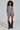 Sporting a stylish look, a person with curly hair is dressed in the OUT OF OFFICE BLAZER ROMPER, an asymmetrical one-sleeve gray blazer dress that exudes business-chic vibes. They pair it effortlessly with black boots accented with chains, standing confidently against a plain background and showcasing their distinctive style.
