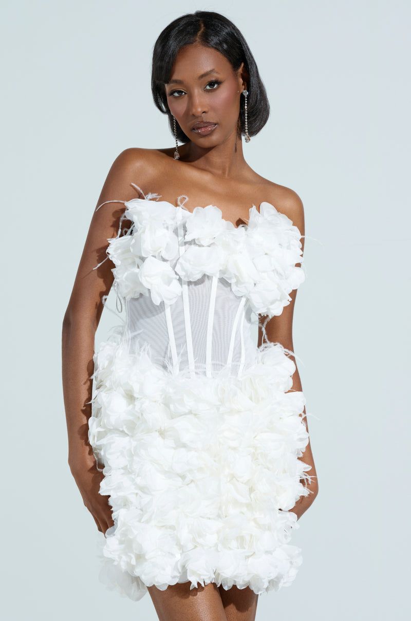 Person in the ON BROADWAY FEATHER AND FLOWER MINI DRESS, featuring a strapless design with a corset-style bodice adorned with large fabric flowers and floral appliques. They have long black hair and are posing against a simple, light-colored background.