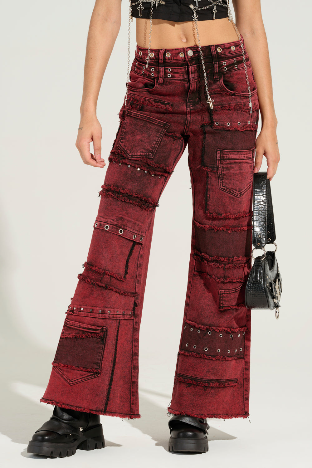 A person is styled in the Grungy Attitude Mineral Wash Red Wide Leg Denim, featuring a patchwork-style design with metal studs and raw hems. They pair it with a black top and chunky black boots, carrying a small black handbag. The image focuses on their outfit, cropped at the shoulders.