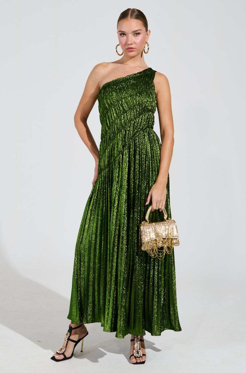 A person poses in the STAY RADIANT METALLIC CHIFFON MAXI DRESS, featuring a shimmering green one-shoulder design. They're holding a gold handbag, wearing black strappy heels and large hoop earrings. The background is plain white.