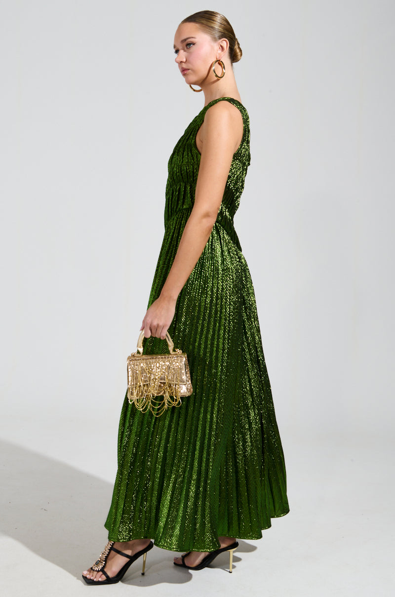 A woman wearing the STAY RADIANT METALLIC CHIFFON MAXI DRESS in shimmering green poses against a plain backdrop. The one-shoulder design complements her gold handbag, while large hoop earrings and black strappy heels complete the look.