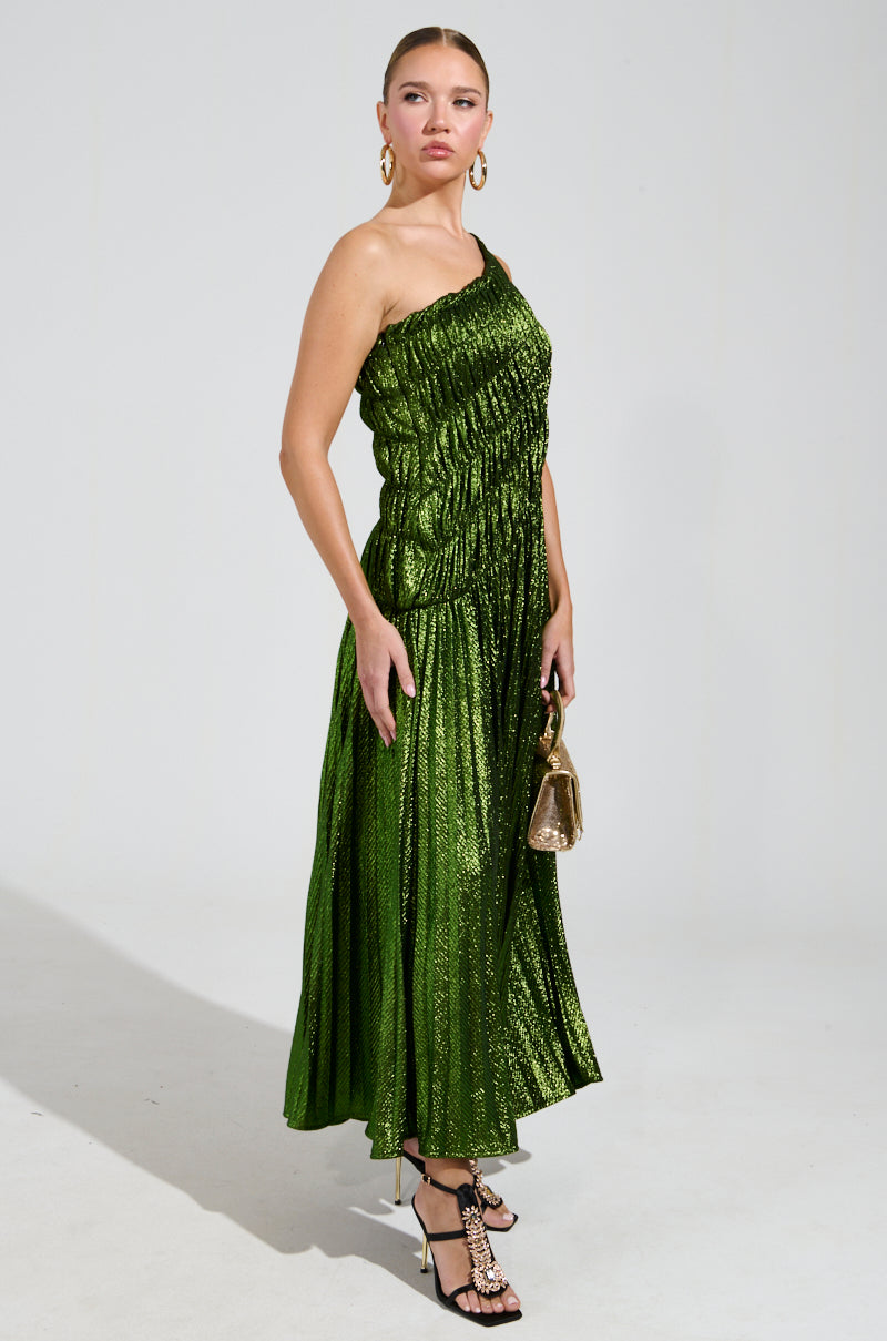 A person stands confidently in the STAY RADIANT METALLIC CHIFFON MAXI DRESS, showcasing a shimmering green look with a ruched bodice. The one-shoulder design features a flowy, pleated style, perfectly complemented by black strappy heels and a small metallic handbag, with the neutral background highlighting the elegant attire.