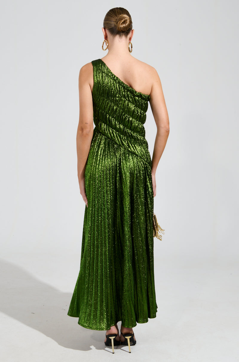 A person stands with their back to the camera, elegantly dressed in the STAY RADIANT METALLIC CHIFFON MAXI DRESS—an exquisite shimmering green gown with pleats and a one-shoulder design; they complement the look with black heels, a neat updo, and gold hoop earrings against a plain background.