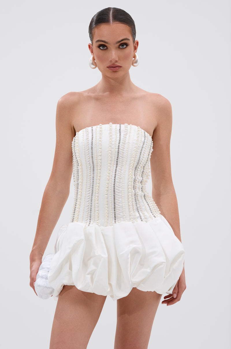 Woman wearing the EXTRA! EXTRA! RHINESTONE MINI DRESS—a strapless white mini with rhinestone details on the fitted bodice and a pleated skirt—holds a white clutch, her hair slicked back and accessorized with large pearl earrings.